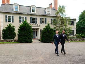 Two men in suits walk outside a large colonial-style white mansion surrounded by lush green trees.