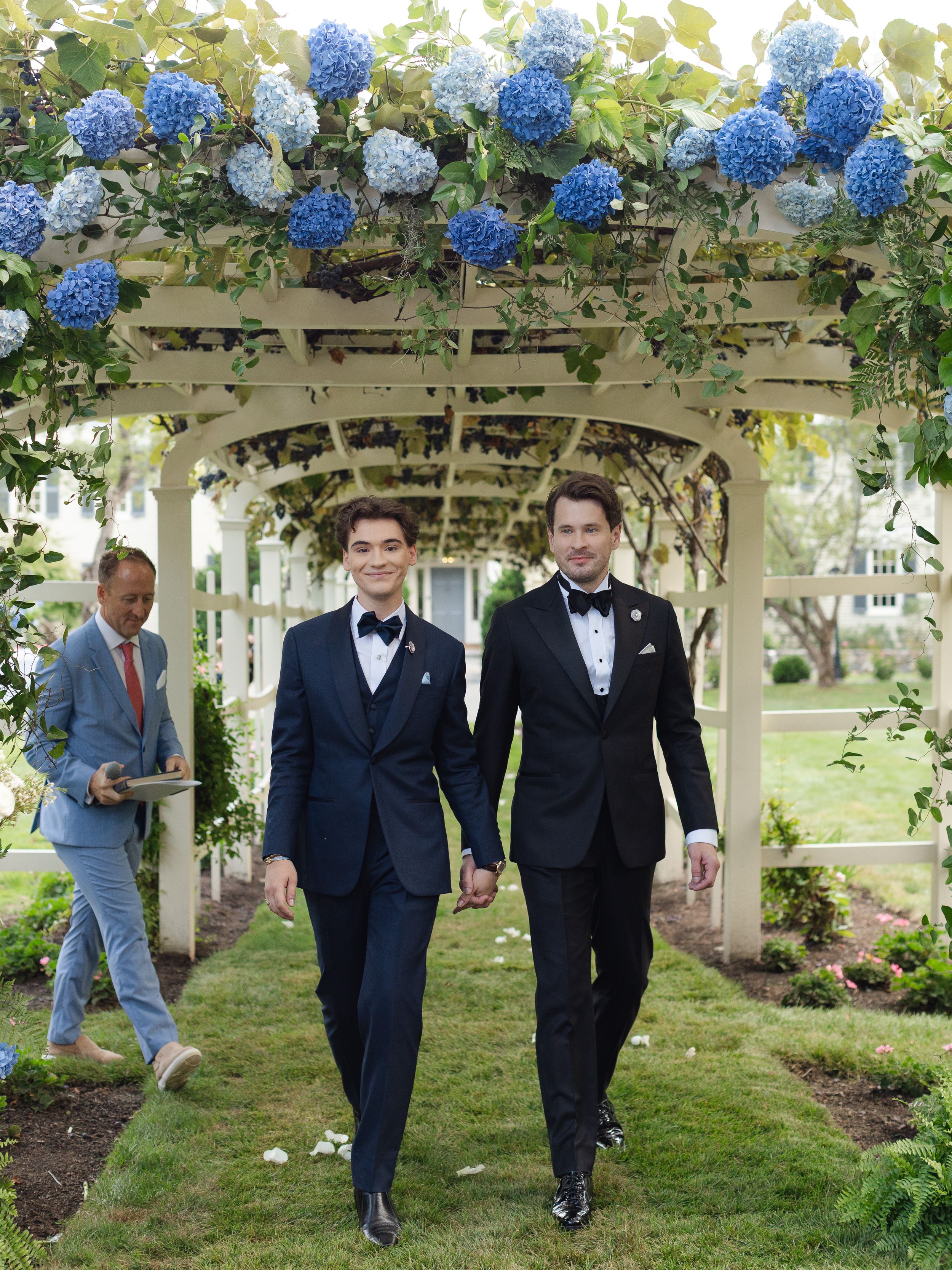 Two grooms in black tuxedos walk hand-in-hand under a white pergola adorned with blue hydrangeas.