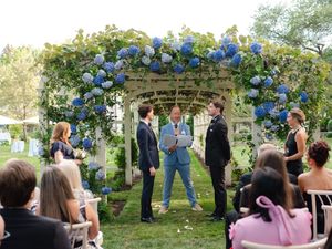 Outdoor same-sex wedding ceremony under white pergola adorned with blue hydrangeas, grooms exchanging vows before guests.