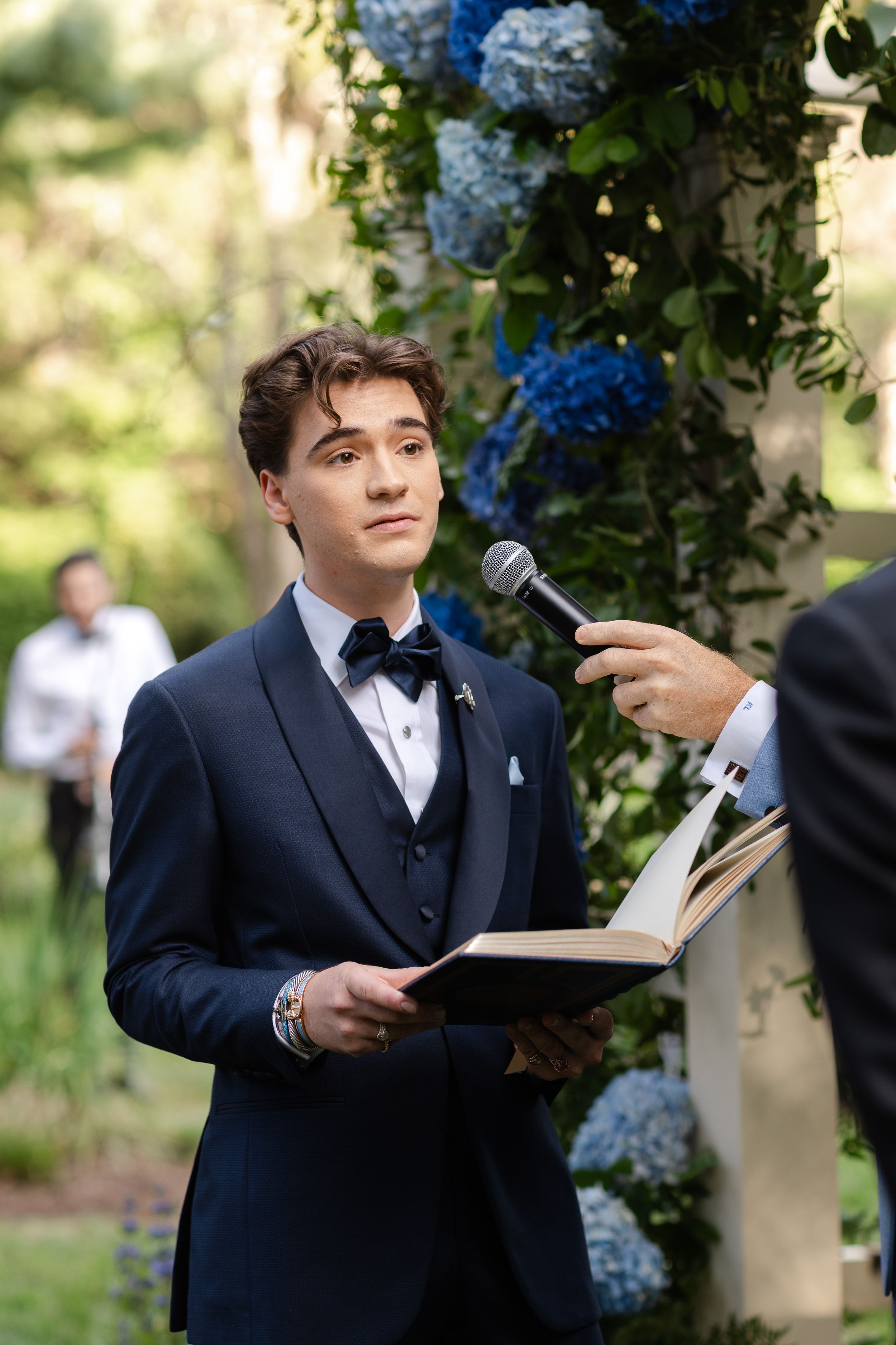 Young man in navy tuxedo holds book at outdoor wedding ceremony with blue hydrangeas.