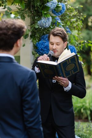 Officiant reading from ceremonial book at outdoor wedding with blue hydrangeas, speaking into microphone.