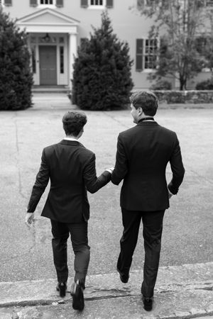 Two men in black suits hold hands walking toward an elegant white building in this black and white photo.