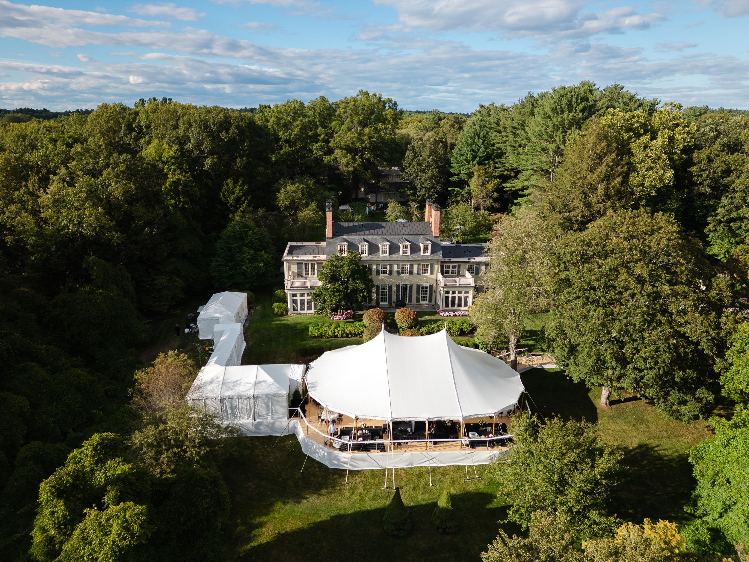 Aerial view of elegant estate mansion with white event tent on lush green grounds surrounded by trees.