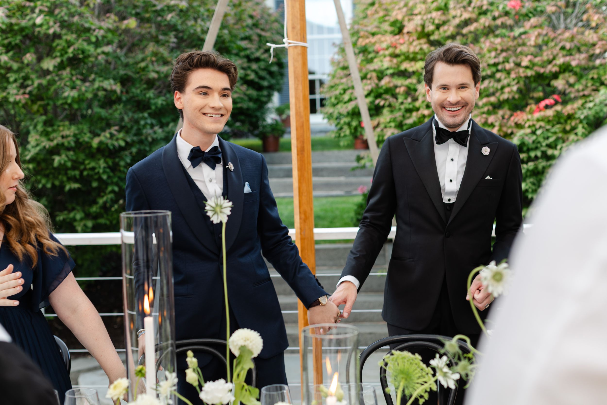 Two grooms smiling and holding hands at an elegant outdoor wedding ceremony with floral decorations.