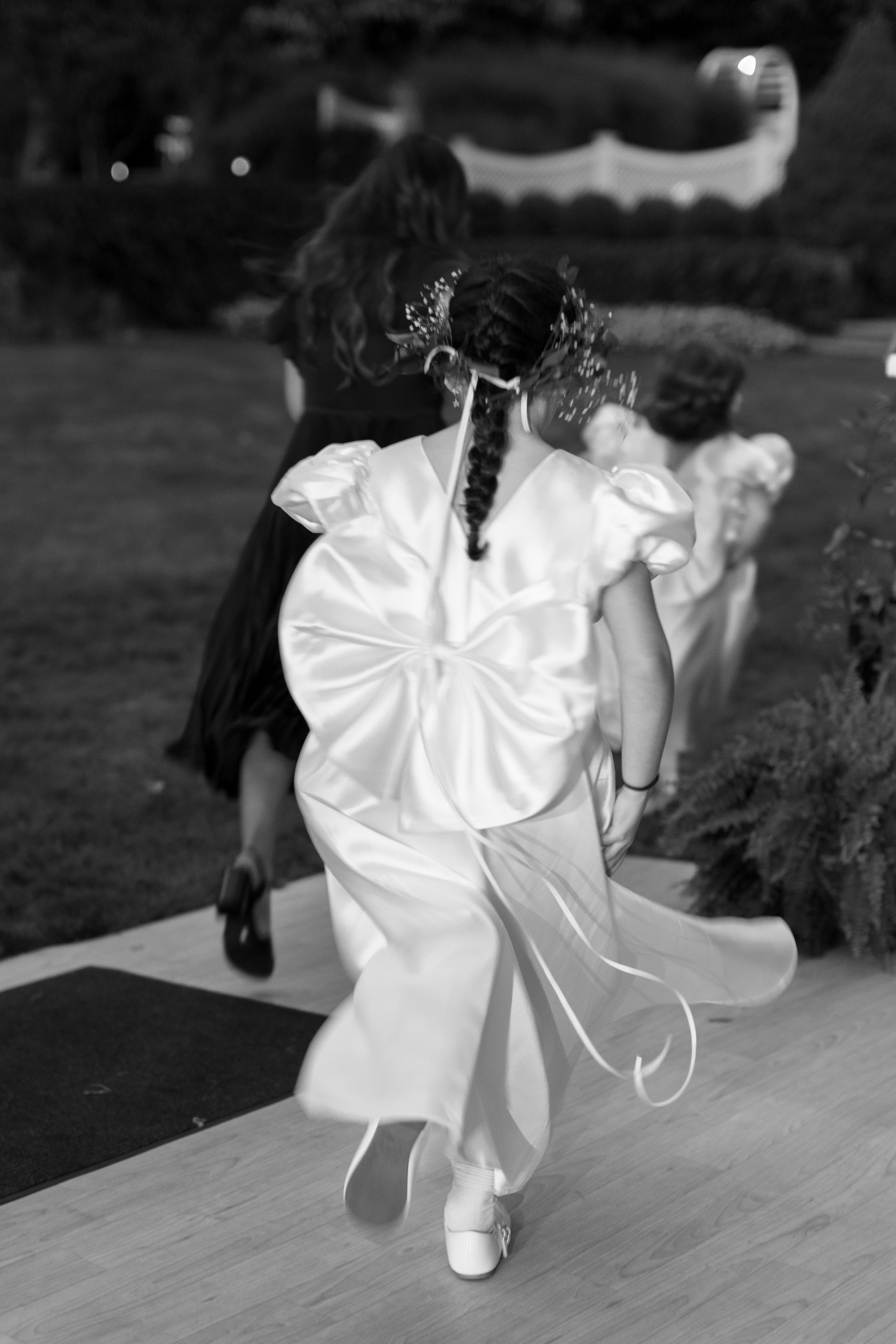 Black and white photo of a flower girl in white dress with bow walking down the aisle at a wedding.
