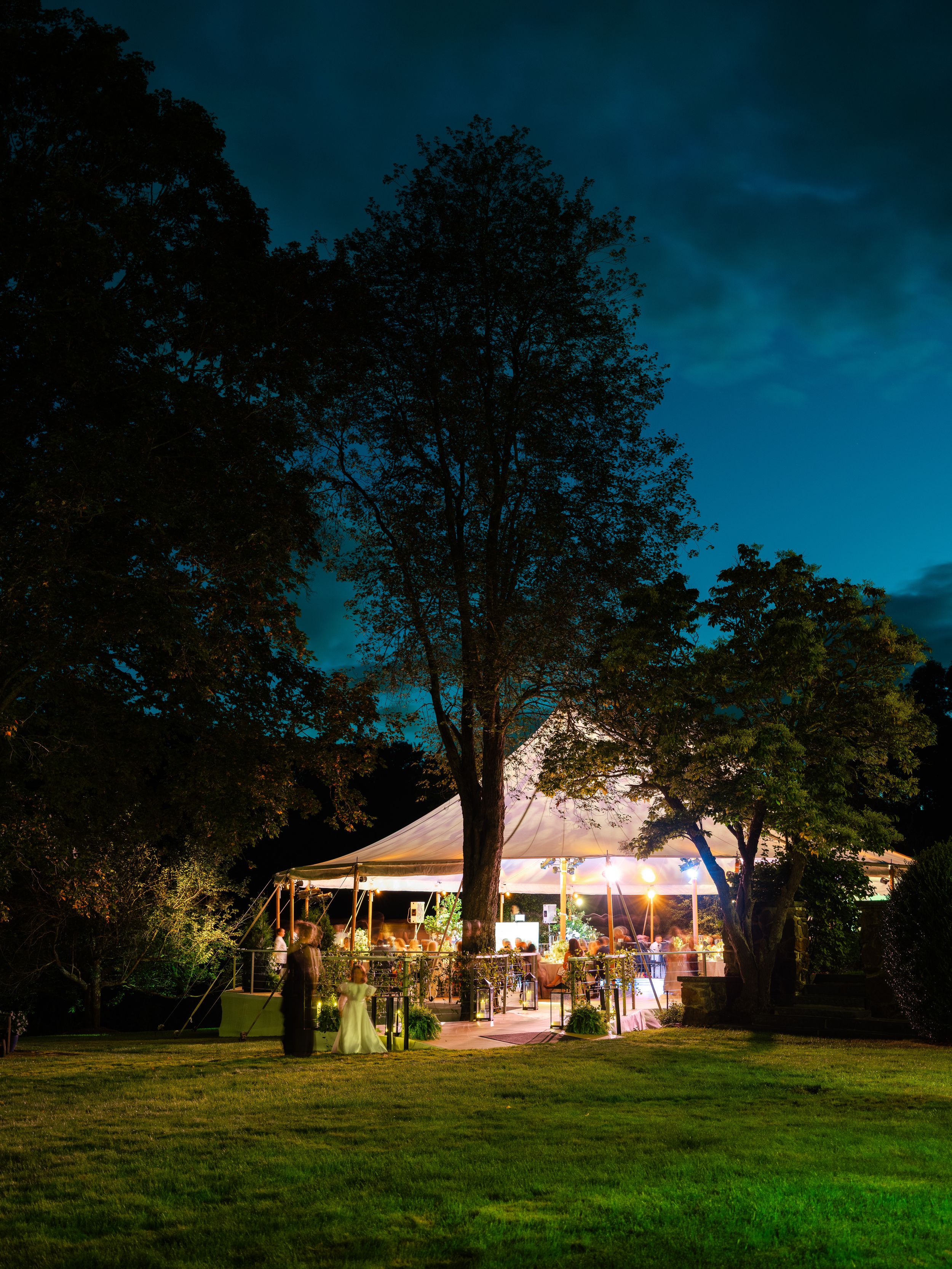 Illuminated outdoor wedding tent glowing warmly at night, surrounded by tall trees under a deep blue evening sky.