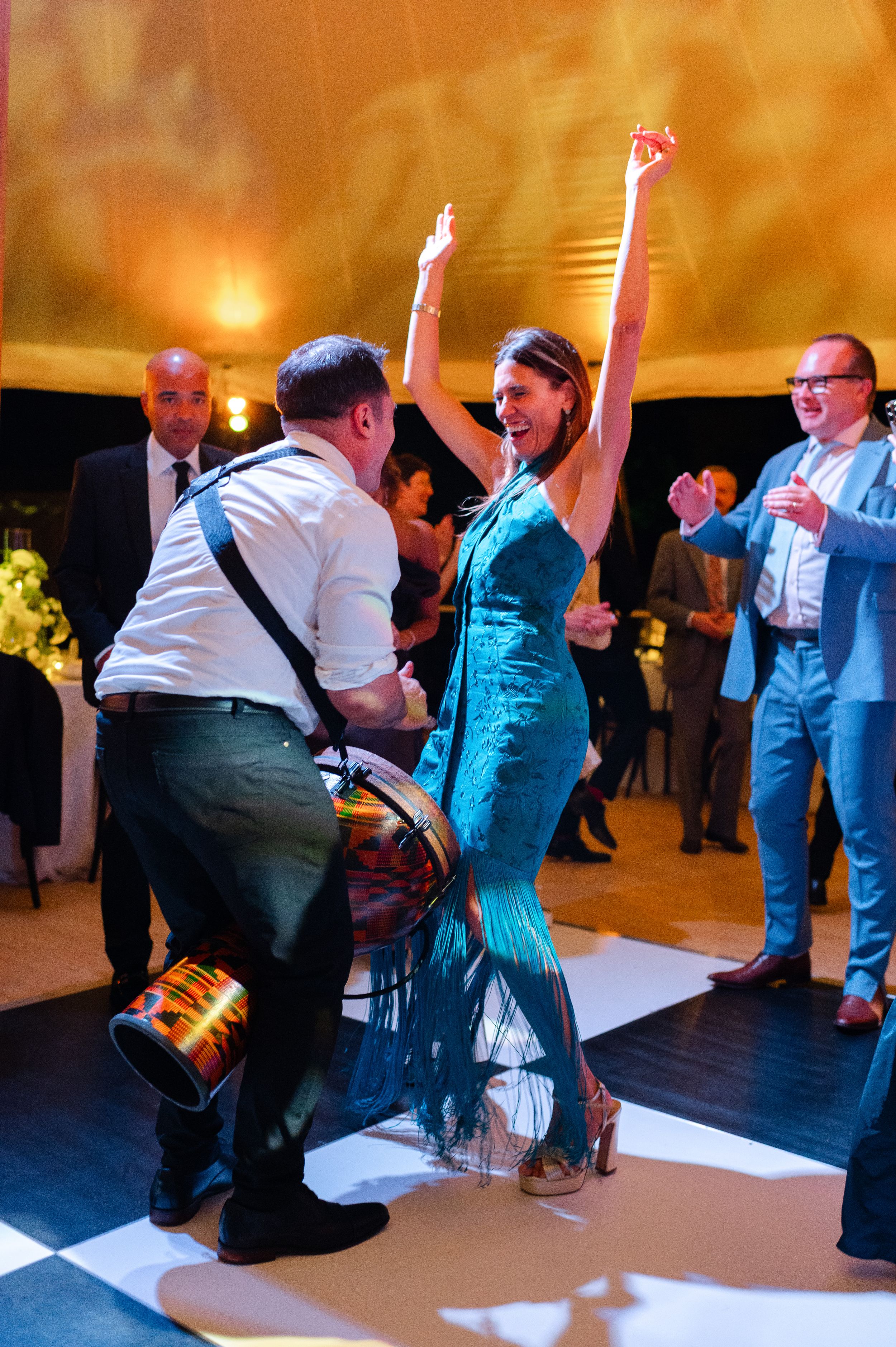 A woman in a teal dress dances joyfully with a drummer at an elegant indoor celebration.