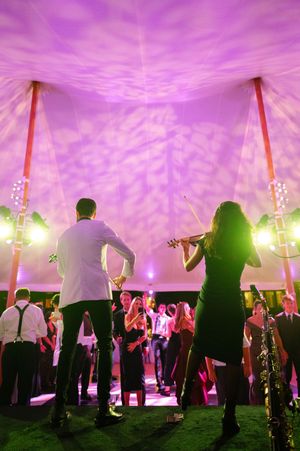 Musicians perform violin and clarinet under a purple-lit tent at an elegant evening event with guests dancing.