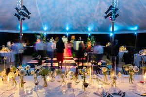 Elegant wedding reception under a white tent with blue lighting, floral centerpieces, and guests dancing on a dance floor.