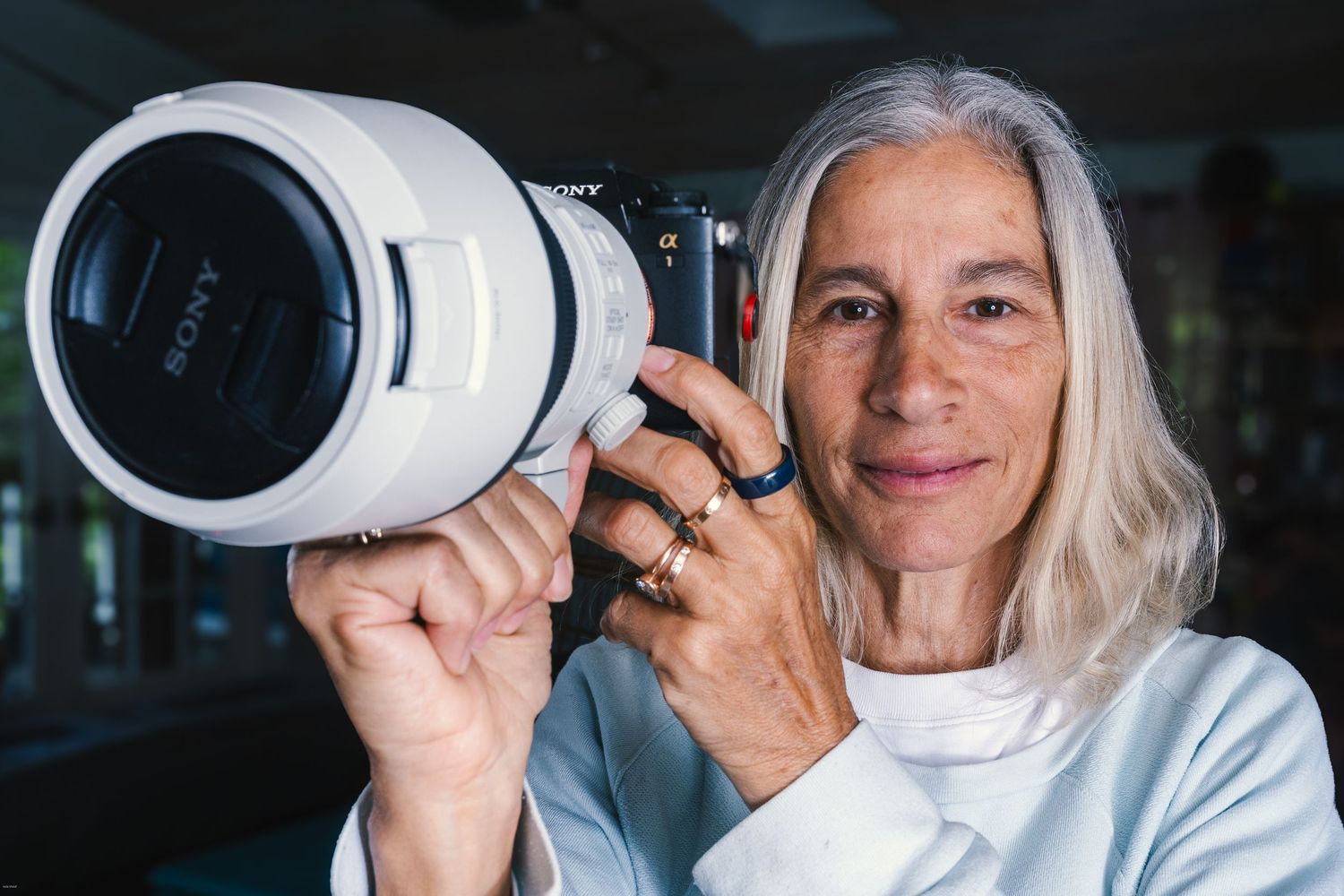 Older woman with gray hair holding a large Sony telephoto lens camera, photographed indoors in soft natural light.