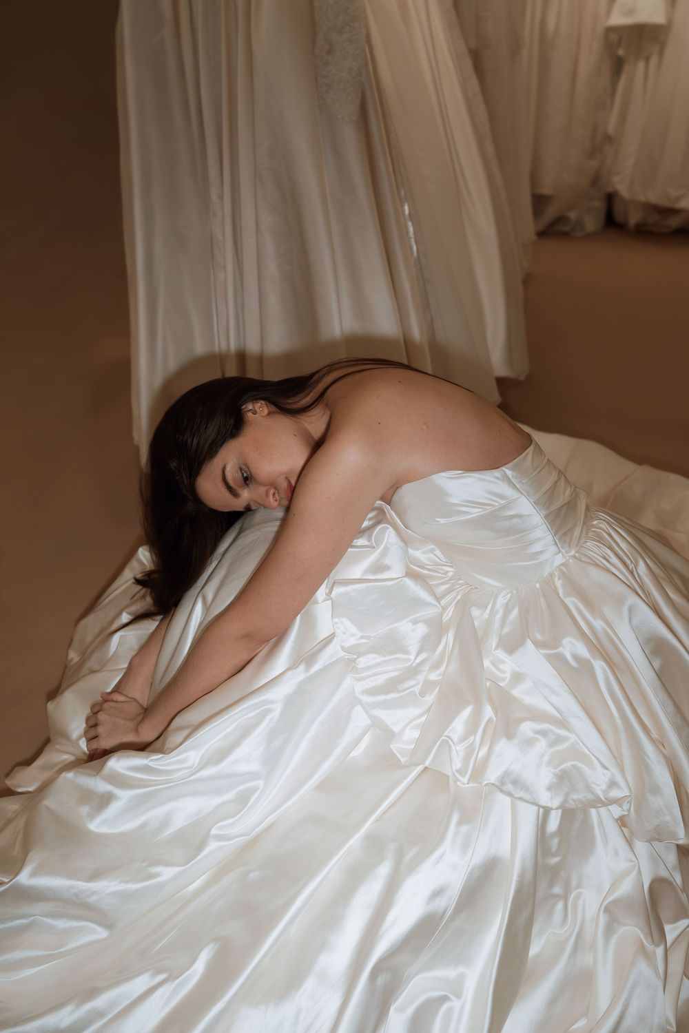 Bride in a strapless white satin ball gown sitting on the floor of a bridal boutique, leaning forward gracefully.