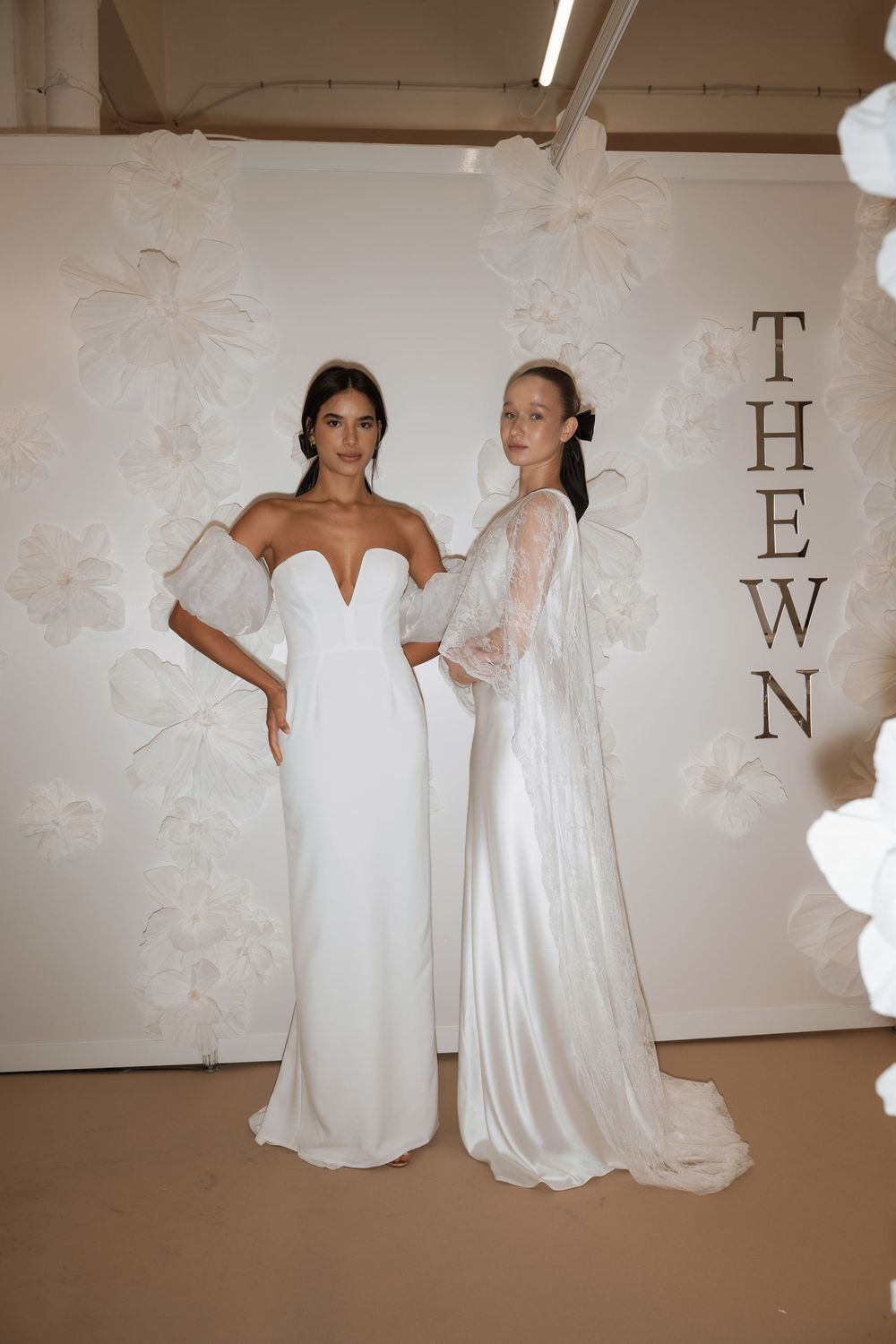 Two models pose in elegant white bridal gowns at The WN bridal fashion showcase, surrounded by floral decor.