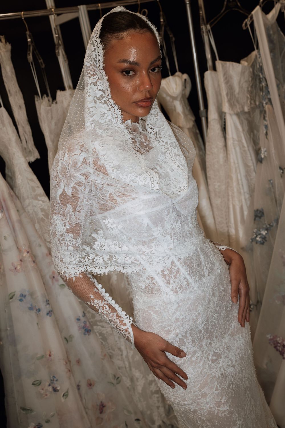 Bride wearing an elegant white lace wedding dress with matching lace veil, surrounded by bridal gowns on a rack.