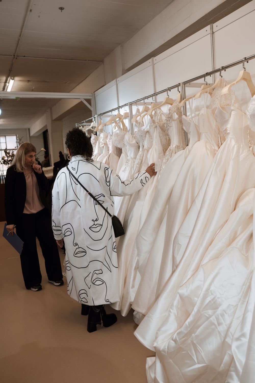Two women browsing elegant white wedding dresses on a rack in a bridal boutique showroom.