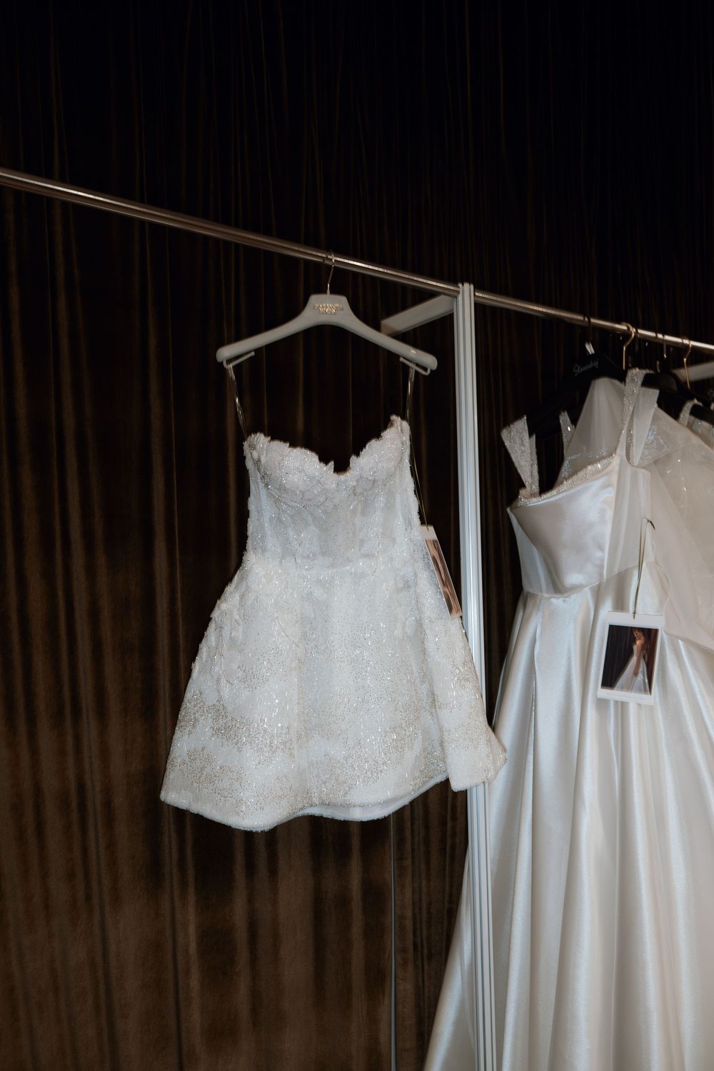 White lace strapless short wedding dress hanging on a rack beside a full-length satin bridal gown.