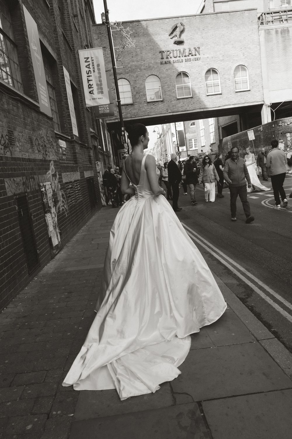 Bride in elegant white gown with train walks Brick Lane street near Truman Brewery, black and white photo.