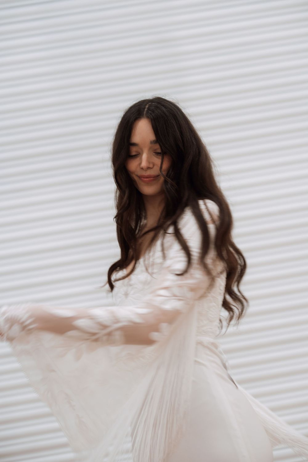 Young woman with long dark hair twirling in a flowing white dress against a white background.