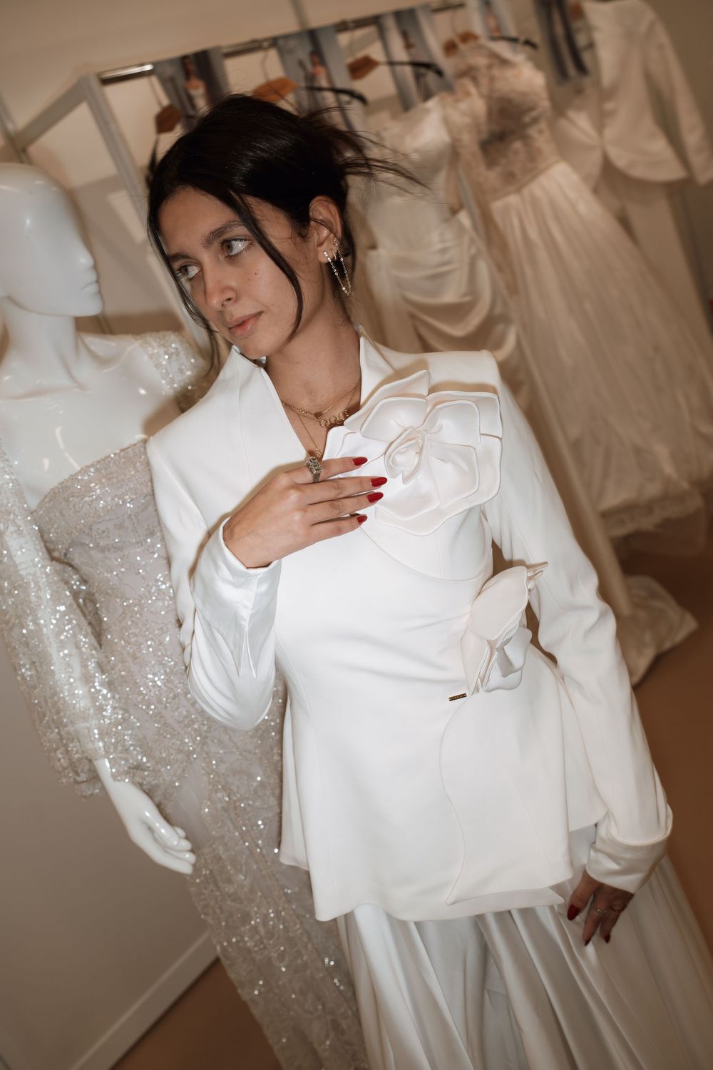 Woman in white blazer with floral detail poses in bridal boutique surrounded by wedding gowns on mannequins.