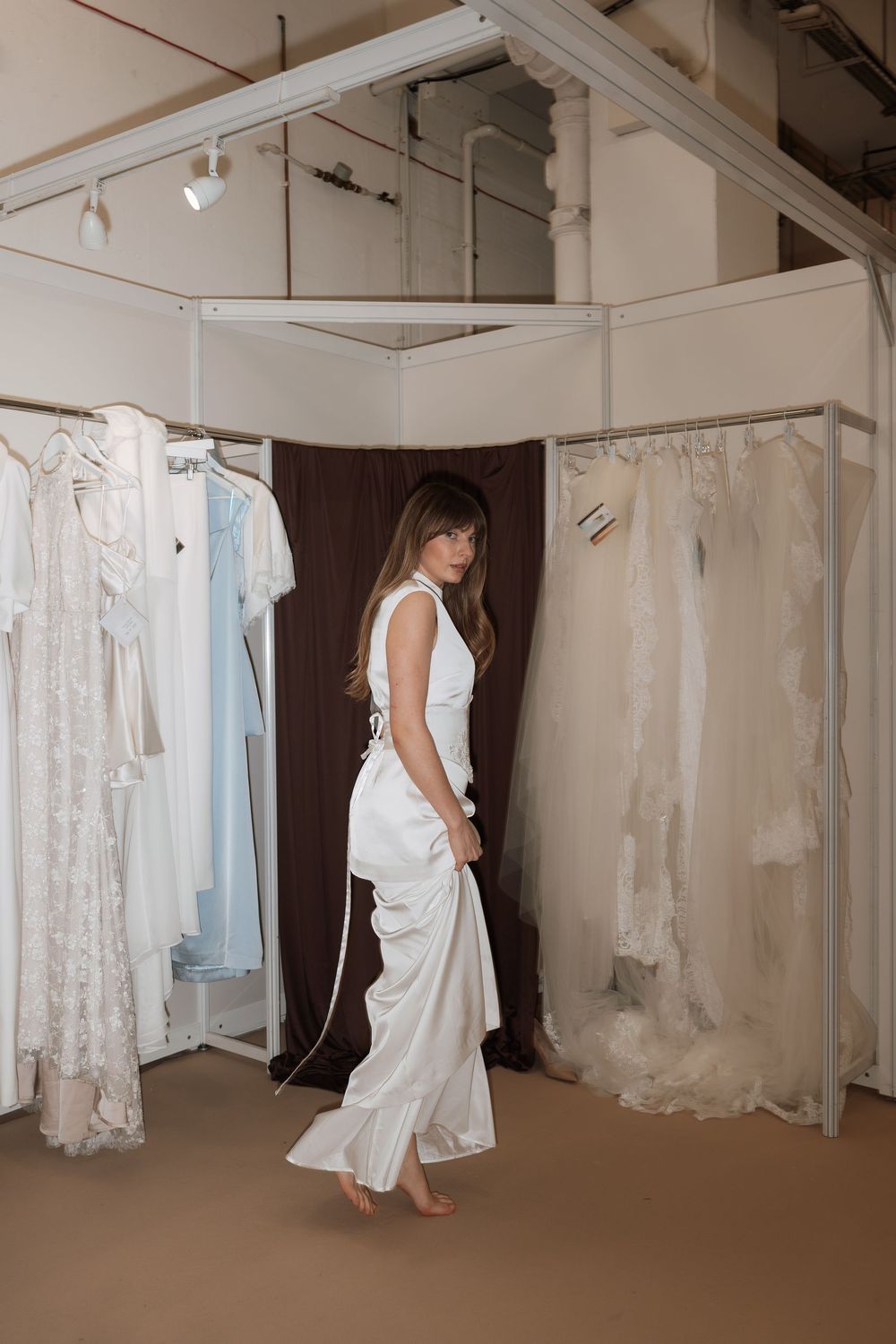 Woman in white satin bridal outfit standing in a bridal boutique fitting room surrounded by wedding dresses.