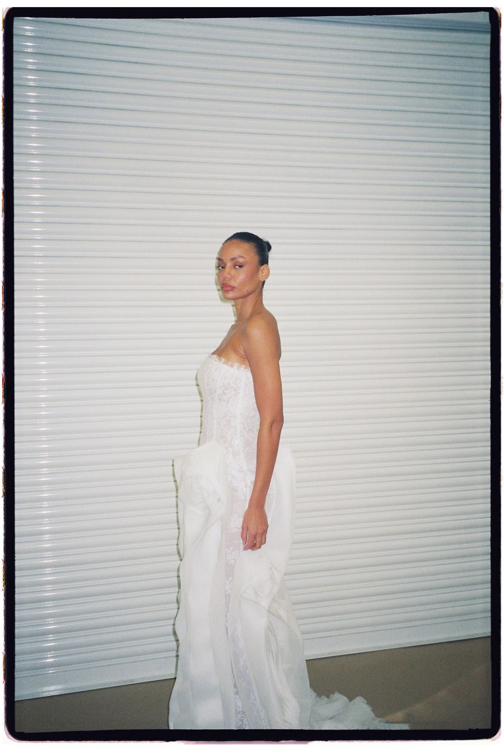 A woman poses in a strapless white bridal gown with ruffled layers against a white slatted background.