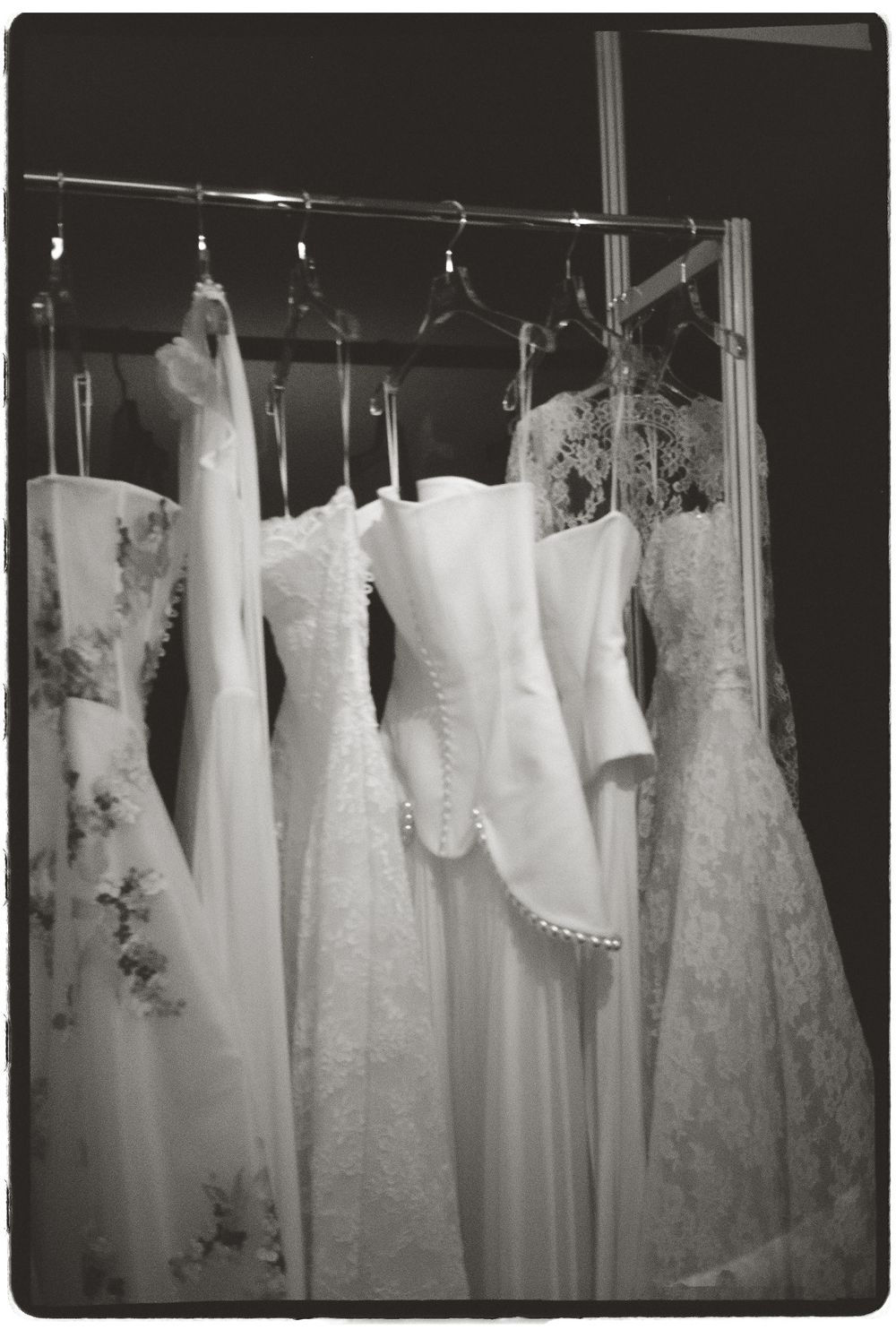 Black and white photo of elegant white wedding gowns hanging on a rack, featuring lace and pearl details.