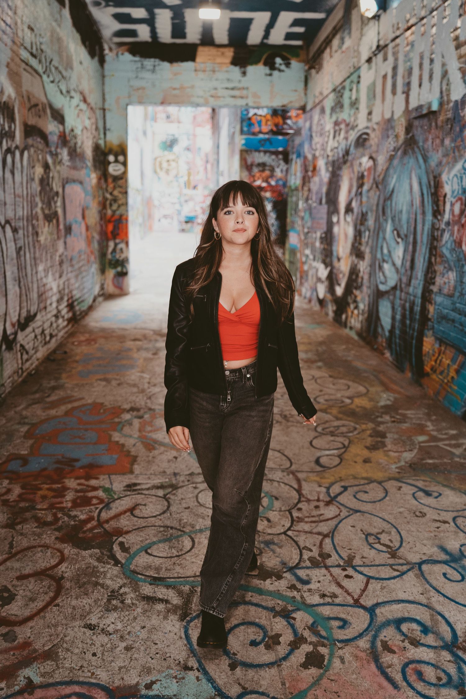 Young woman in red crop top and black blazer walks through graffiti-covered urban alley tunnel.