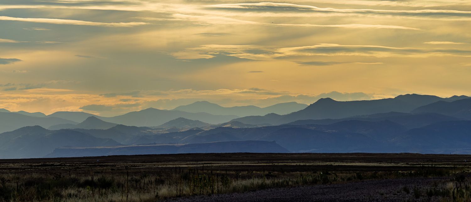 Panoramic sunset view of layered mountain silhouettes under golden cloudy skies over a vast open plain.