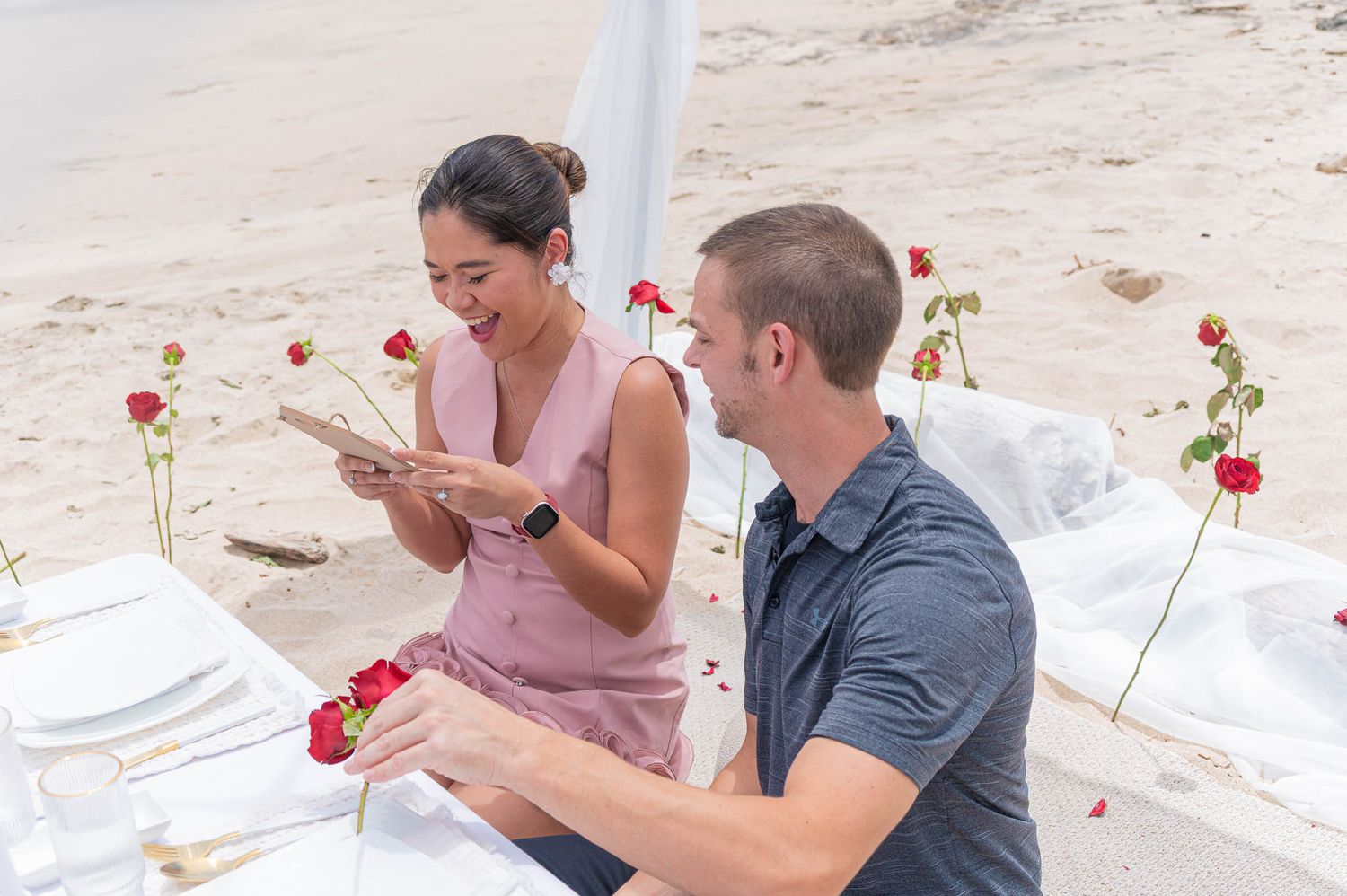 Couple enjoying a romantic beach dinner with red roses, woman reading a note while man smiles at a white-draped table.