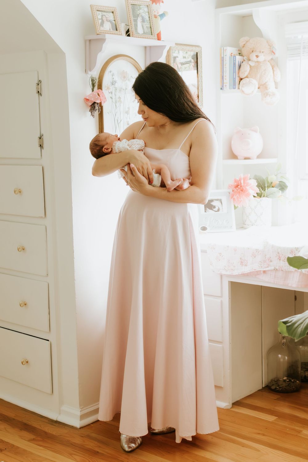 A mother in a blush pink gown holds her newborn baby in a soft, elegantly decorated nursery room.
