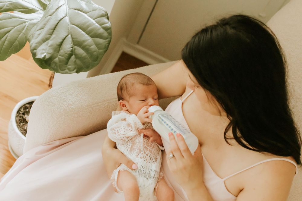 A mother bottle-feeding her newborn baby while sitting on a bed, with a green plant in the background.