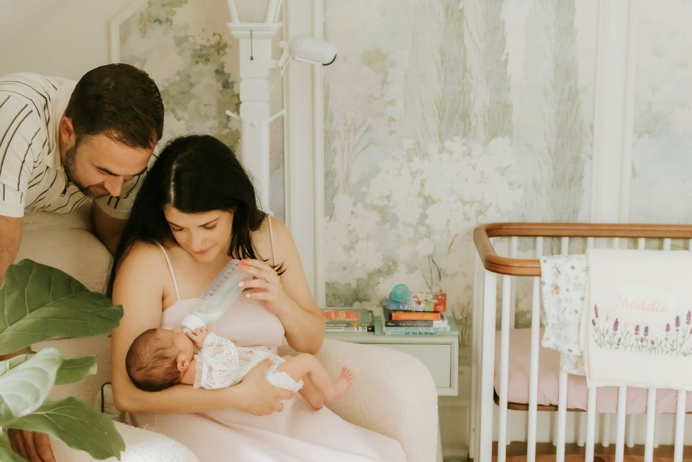 New parents admire their newborn baby in a softly decorated nursery with floral wallpaper and wooden crib.
