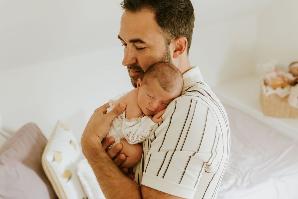 A loving father gently cradles his sleeping newborn baby against his chest in a bright, cozy nursery setting.