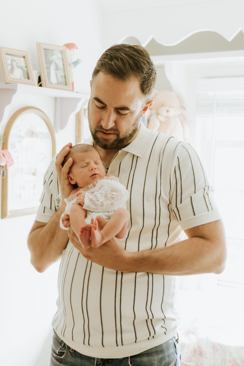 A bearded father tenderly cradles his sleeping newborn baby in a softly lit nursery room.