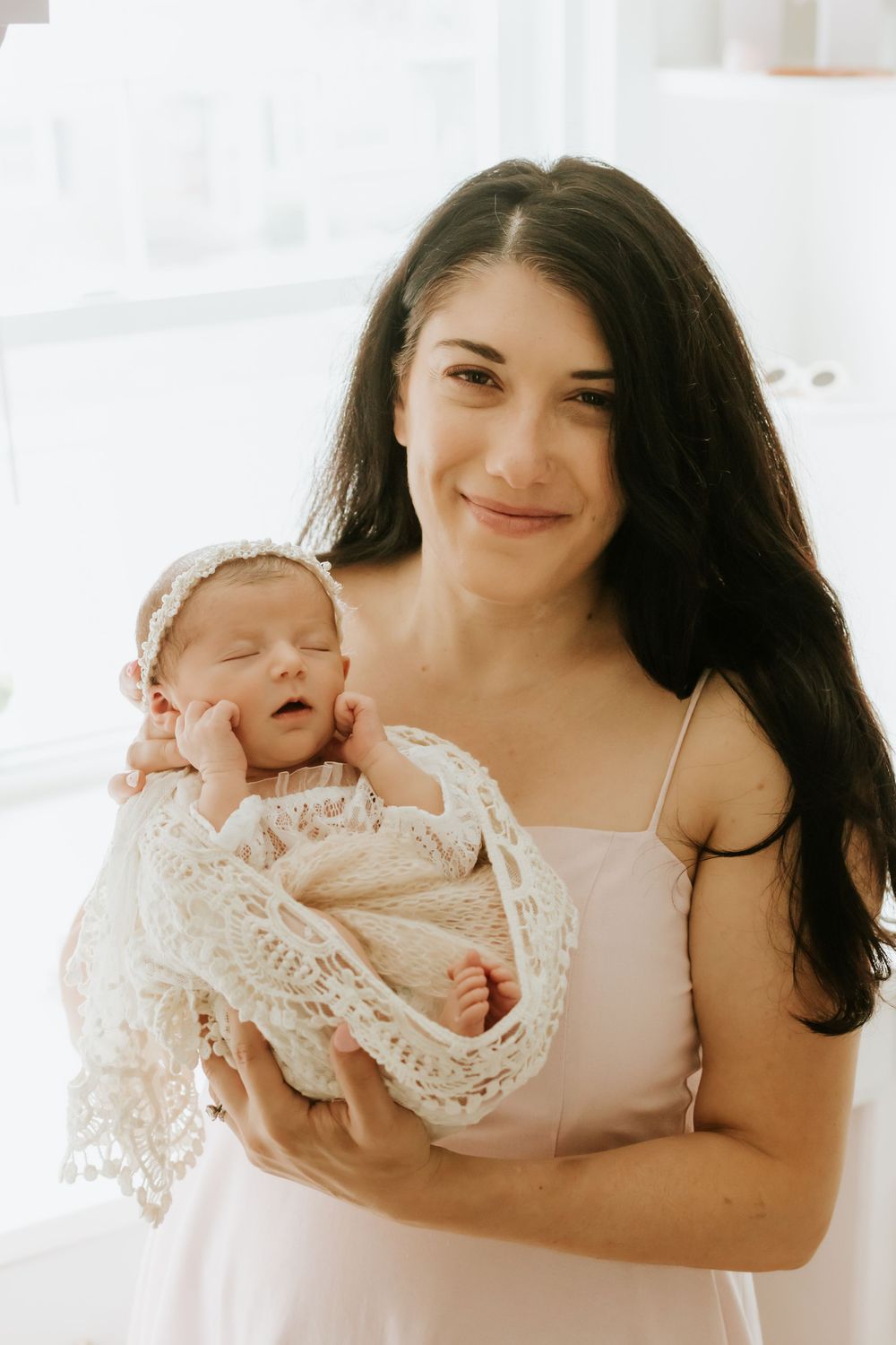 A smiling dark-haired woman in a pink dress holds a sleeping newborn wrapped in a lace blanket and bonnet.