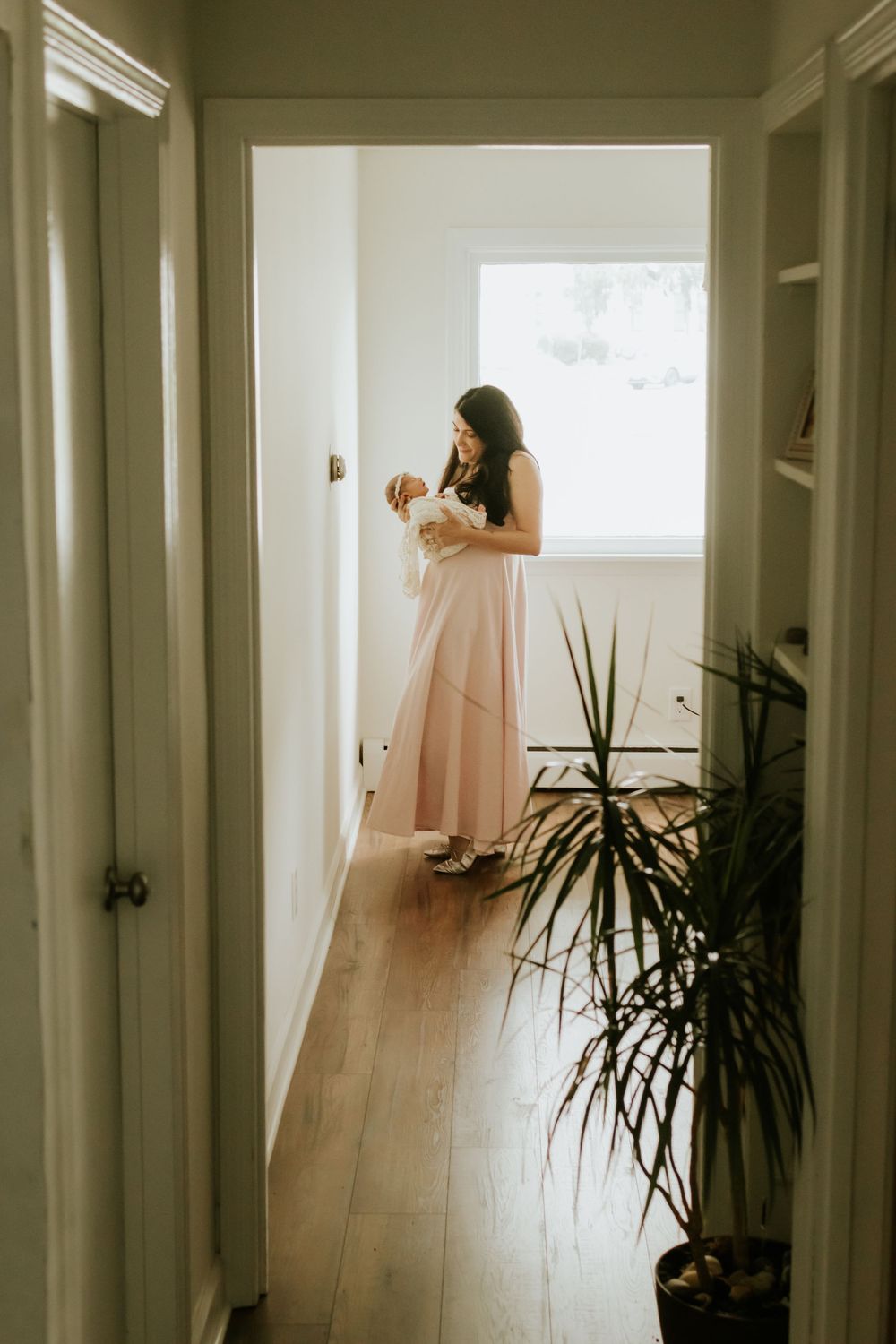 A woman in a pink maxi dress holds a newborn baby in a sunlit hallway with hardwood floors and a green plant.