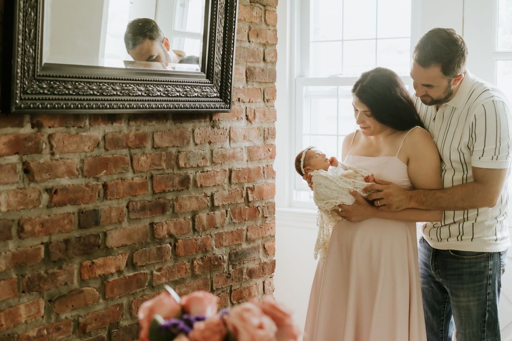 New parents tenderly hold their newborn baby in a bright room with exposed brick walls and a decorative mirror.