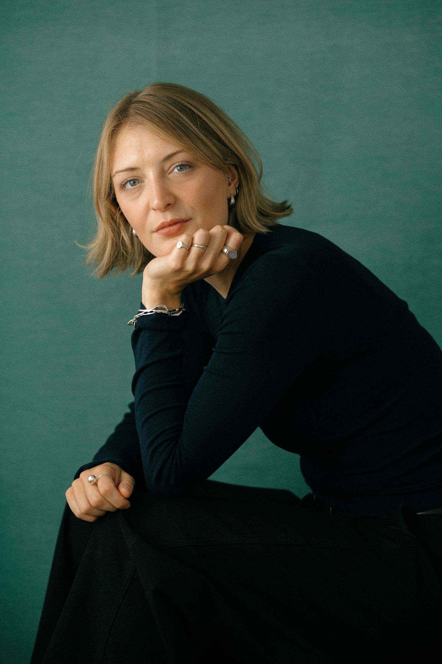 Woman in black top sits pensively against teal background, resting chin on hand, wearing silver jewelry.