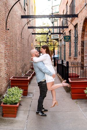 A man lifts a woman in a romantic embrace in a charming brick alley with potted plants and vintage lanterns.