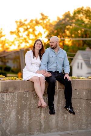 A couple sits together on a stone wall at golden hour, smiling and holding hands outdoors.