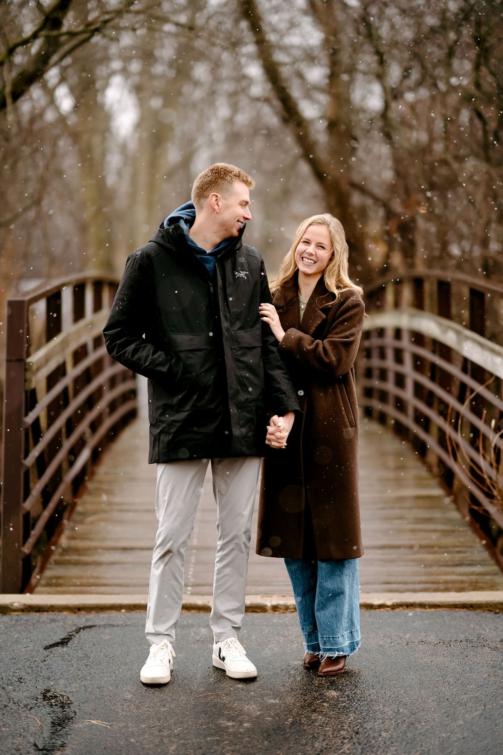 A couple stands together on a wooden bridge during a moody winter day, wearing warm coats and sharing a moment.