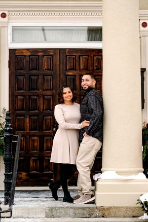 Couple smiling and posing together in front of elegant wooden doors on a snowy day.