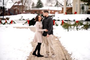 Couple holding hands on a snowy brick path with festive holiday wreaths and red bows in the background.