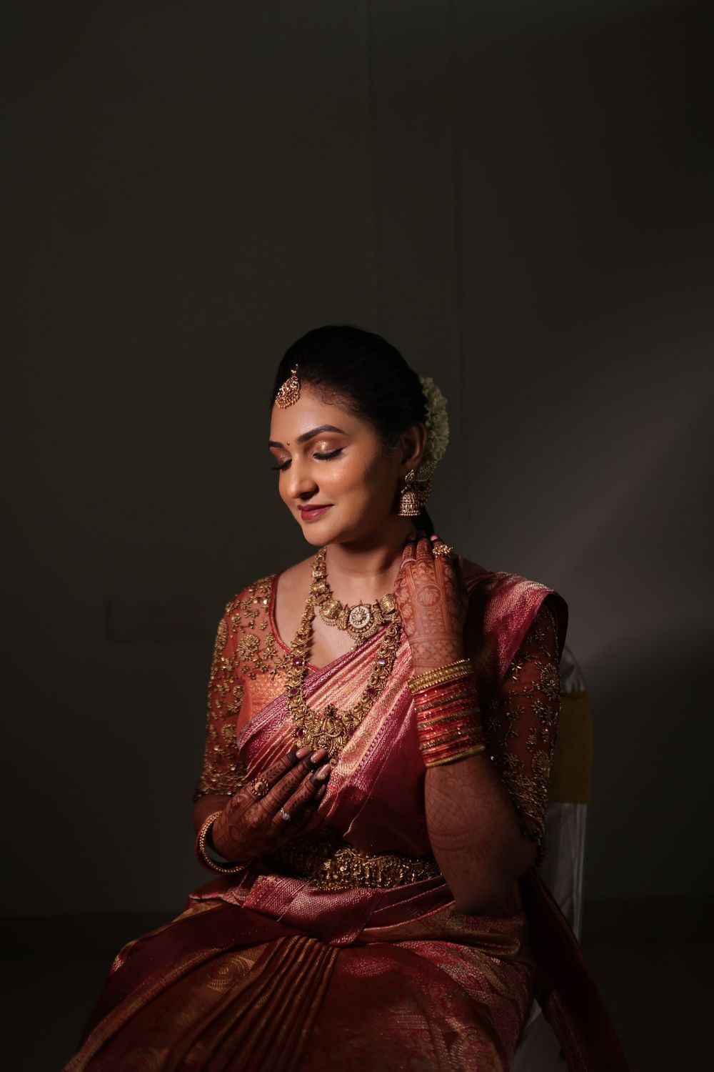 South Indian bride in pink silk saree with gold jewelry and floral hair accessories, smiling gracefully in studio portrait.