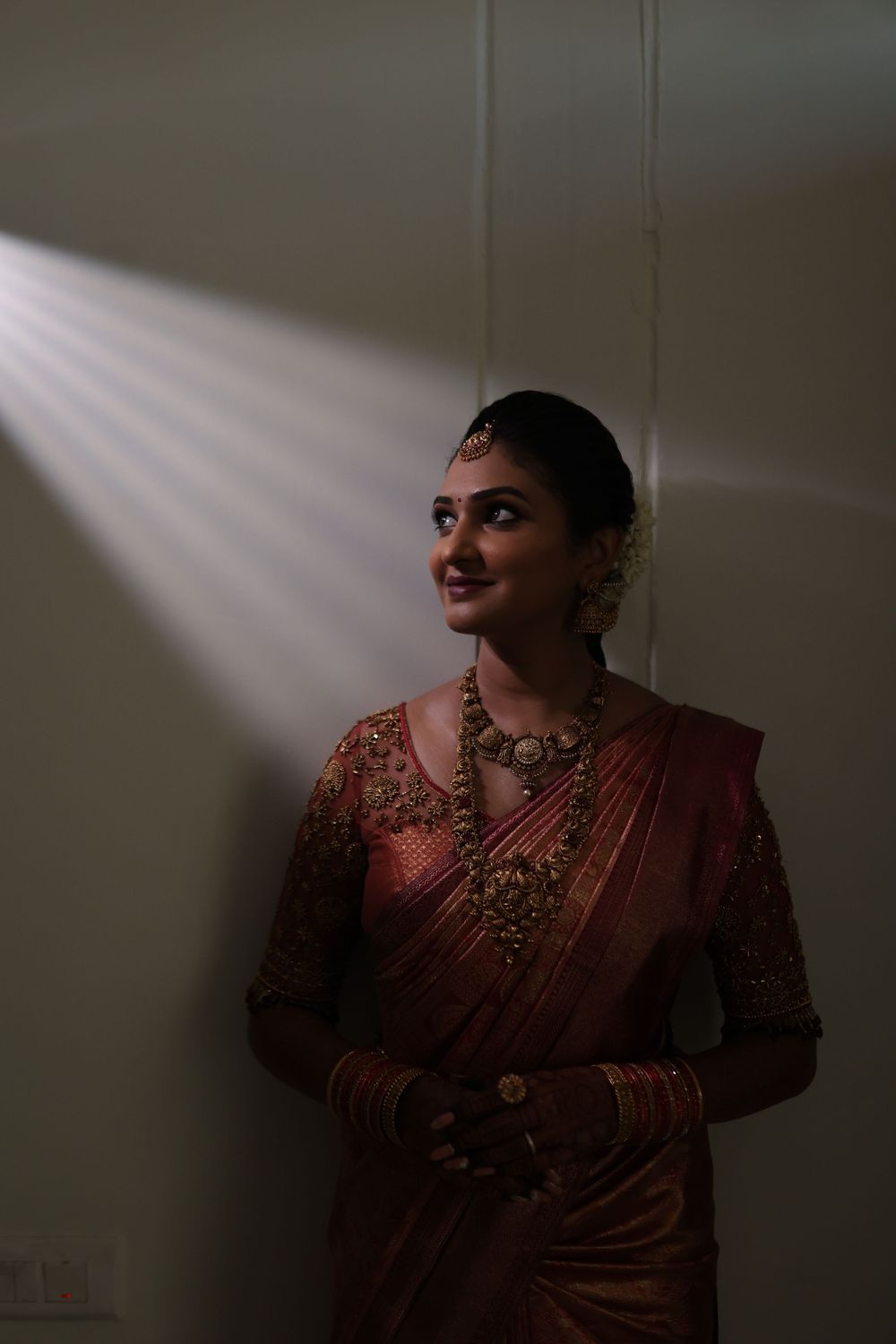 A bride in a red silk saree with gold jewelry stands in dramatic light against a wall, smiling softly.