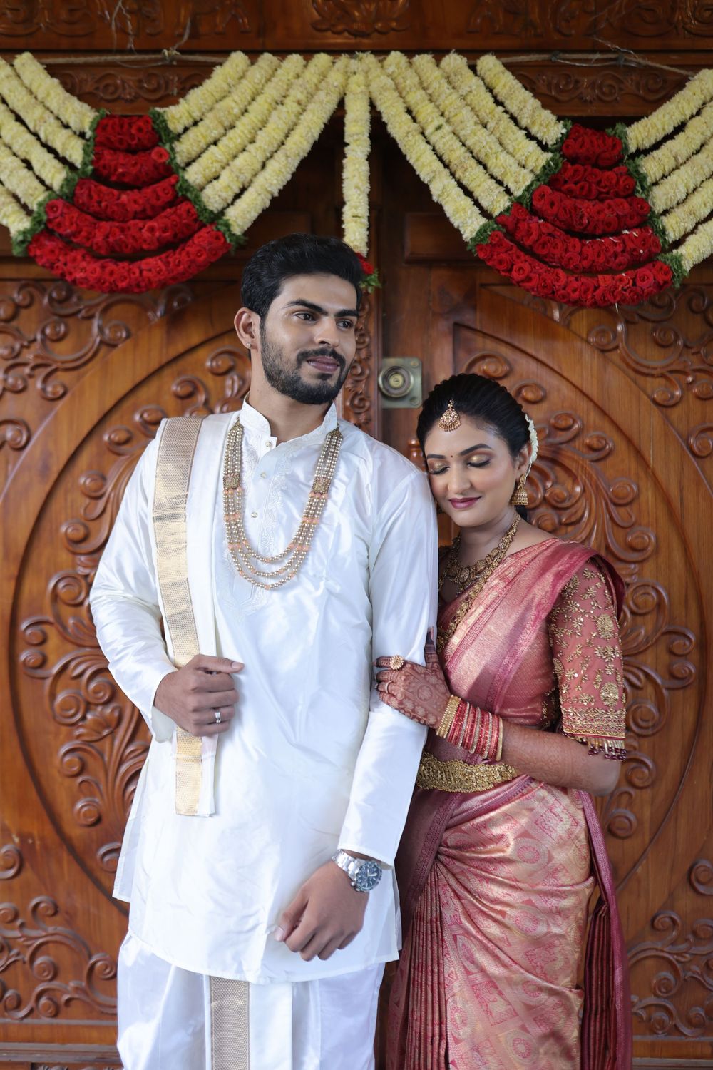 South Indian couple in traditional wedding attire posing before ornate wooden door with floral garlands.