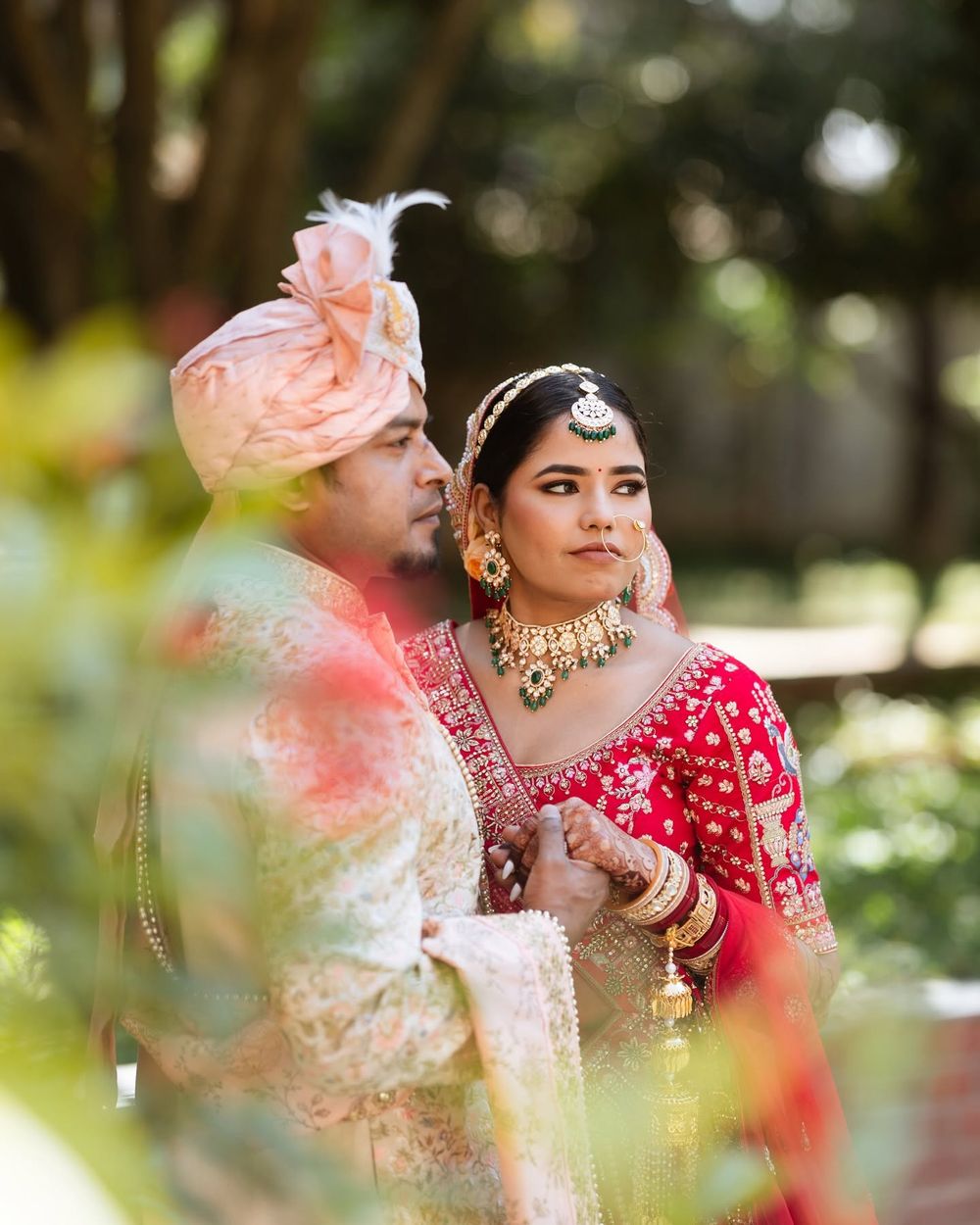 Indian bride in red lehenga and groom in ivory sherwani with pink turban pose together at outdoor wedding ceremony.