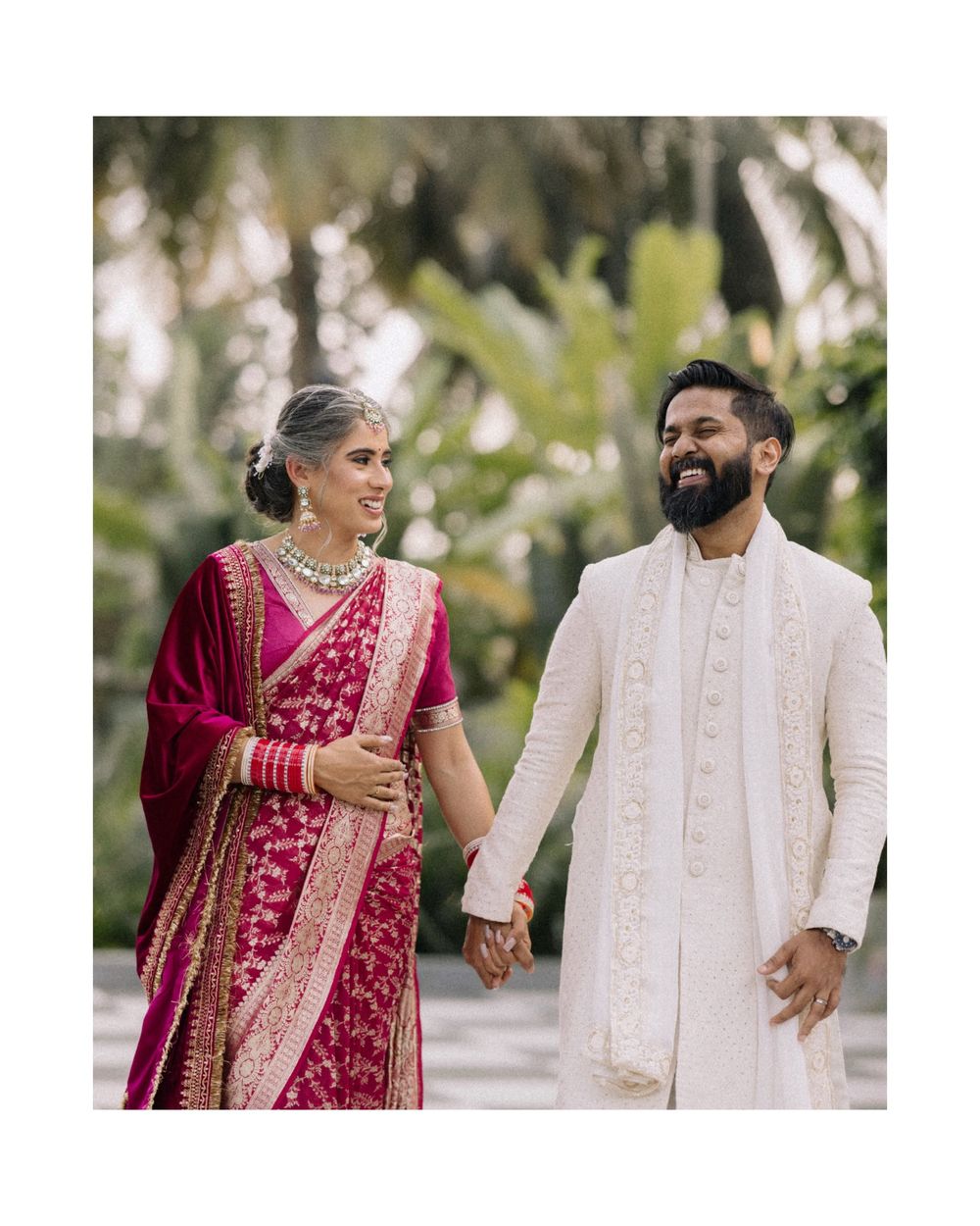 Indian couple in traditional wedding attire walk hand-in-hand, bride in red saree and groom in white sherwani.