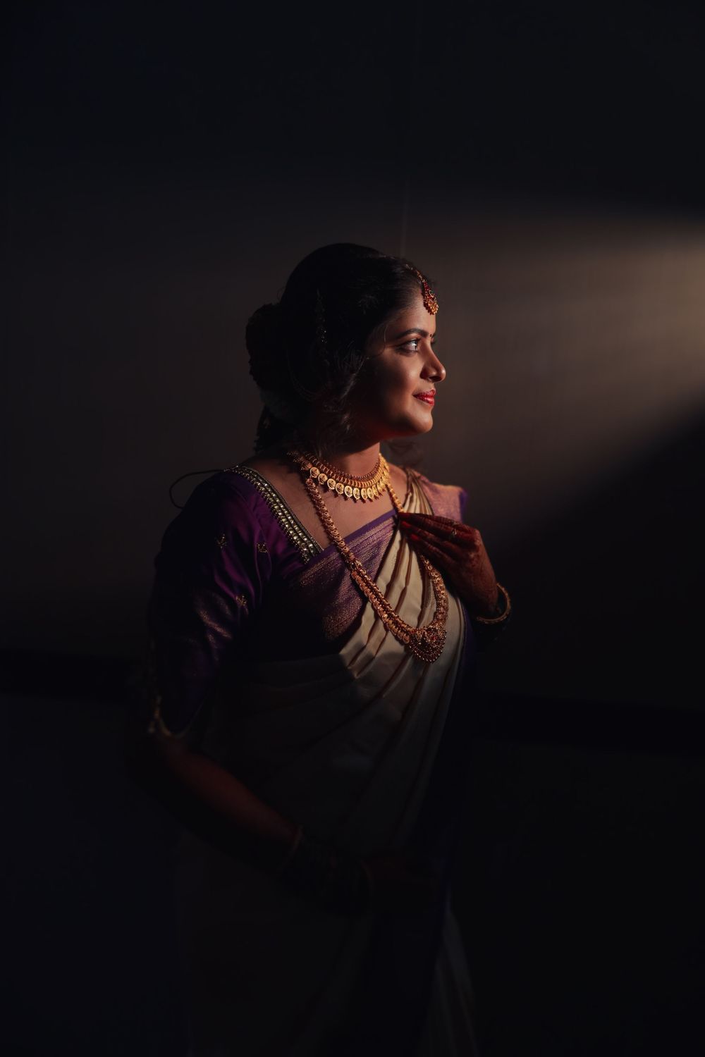 Elegant Indian bride in purple and white saree posed dramatically in moody studio lighting with gold jewelry.