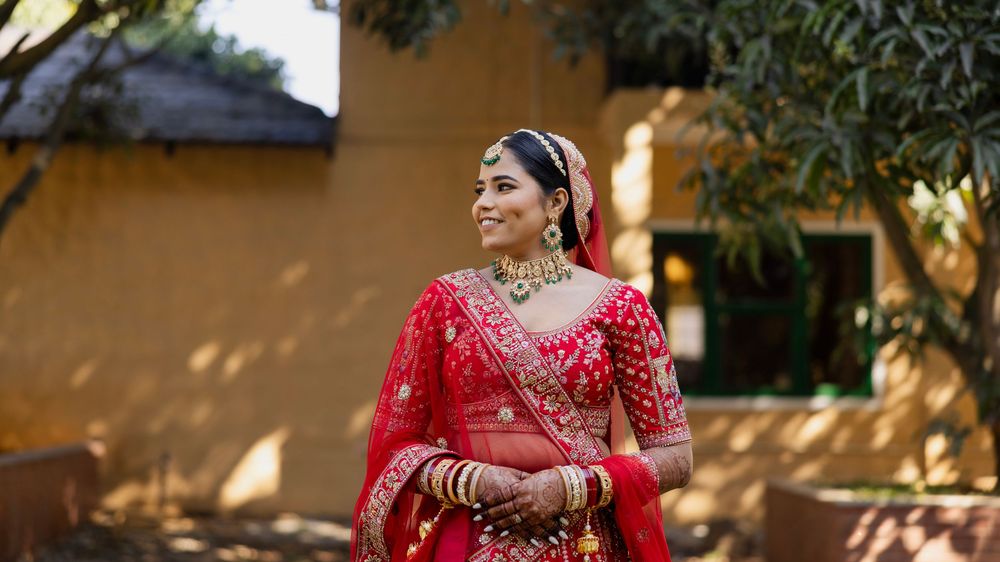 Indian bride in red lehenga with gold jewelry poses outdoors in traditional Rajasthani bridal attire.