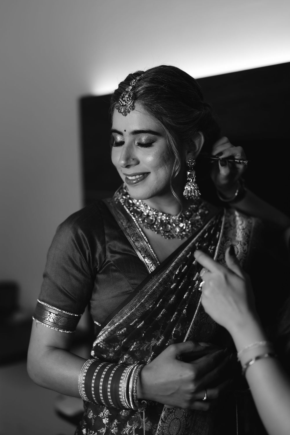 Black and white photo of a Indian bride in saree and jewelry being dressed, smiling softly.